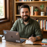 A man sitting at a wooden desk, smiling and writing in a notebook, with a laptop and books in the background.